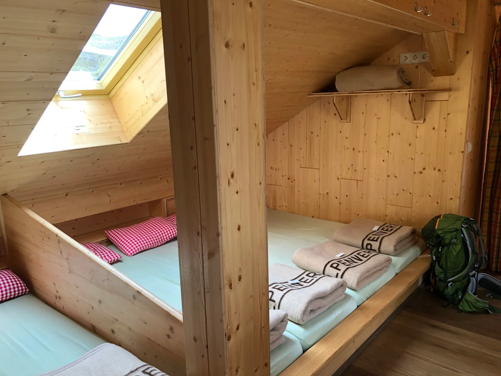 Matratzenlager unter dem Dach, Holzverschläge mit Decken und Kissen, daneben ein Rucksack